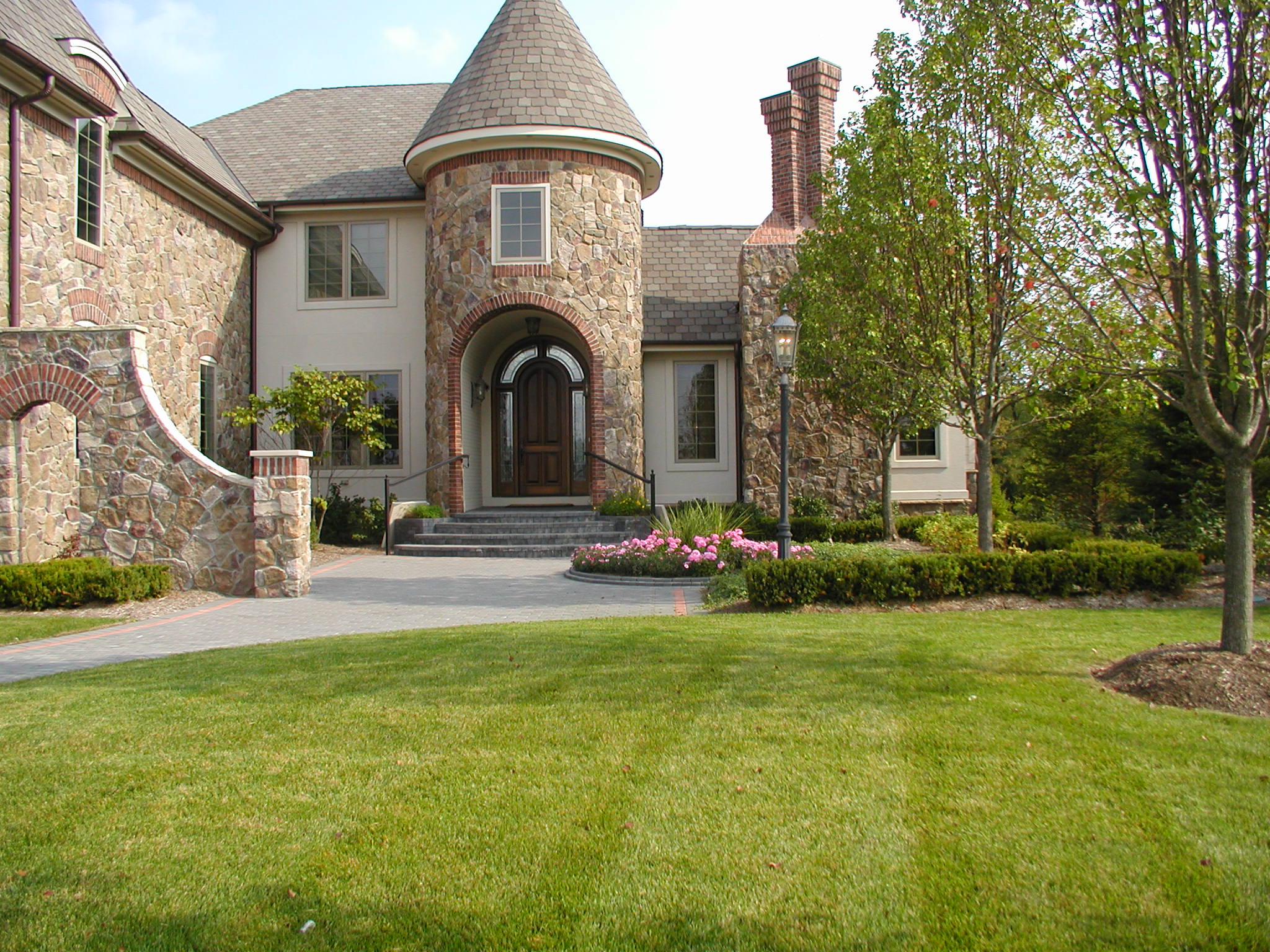 A picturesque stone house with a turret entrance, lush green lawn, and surrounding trees on a sunny day. Garden features vibrant pink flowers.