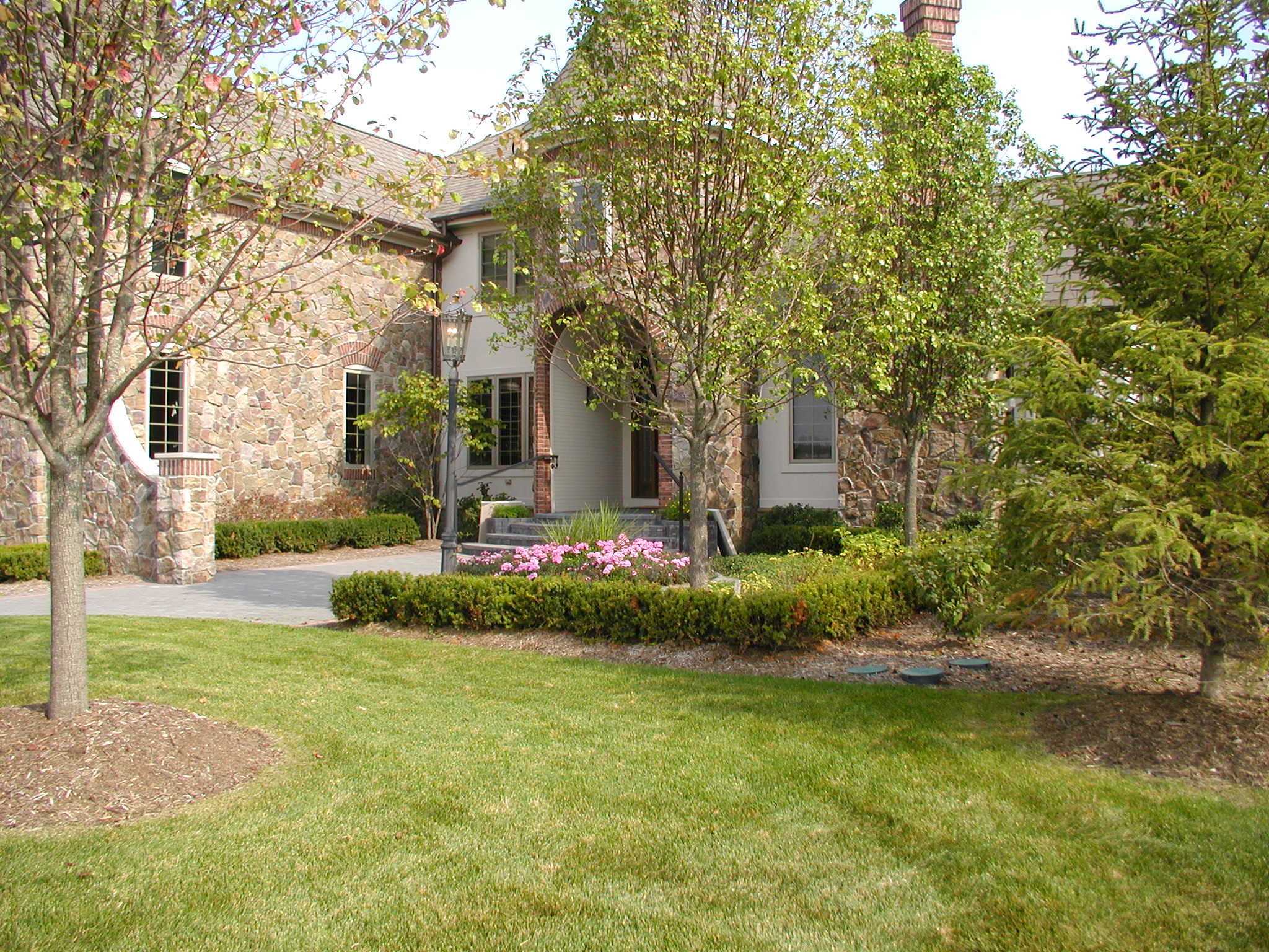A stone house surrounded by lush greenery and blooming flowers, featuring a pathway lined with trimmed bushes and a traditional outdoor lamp.