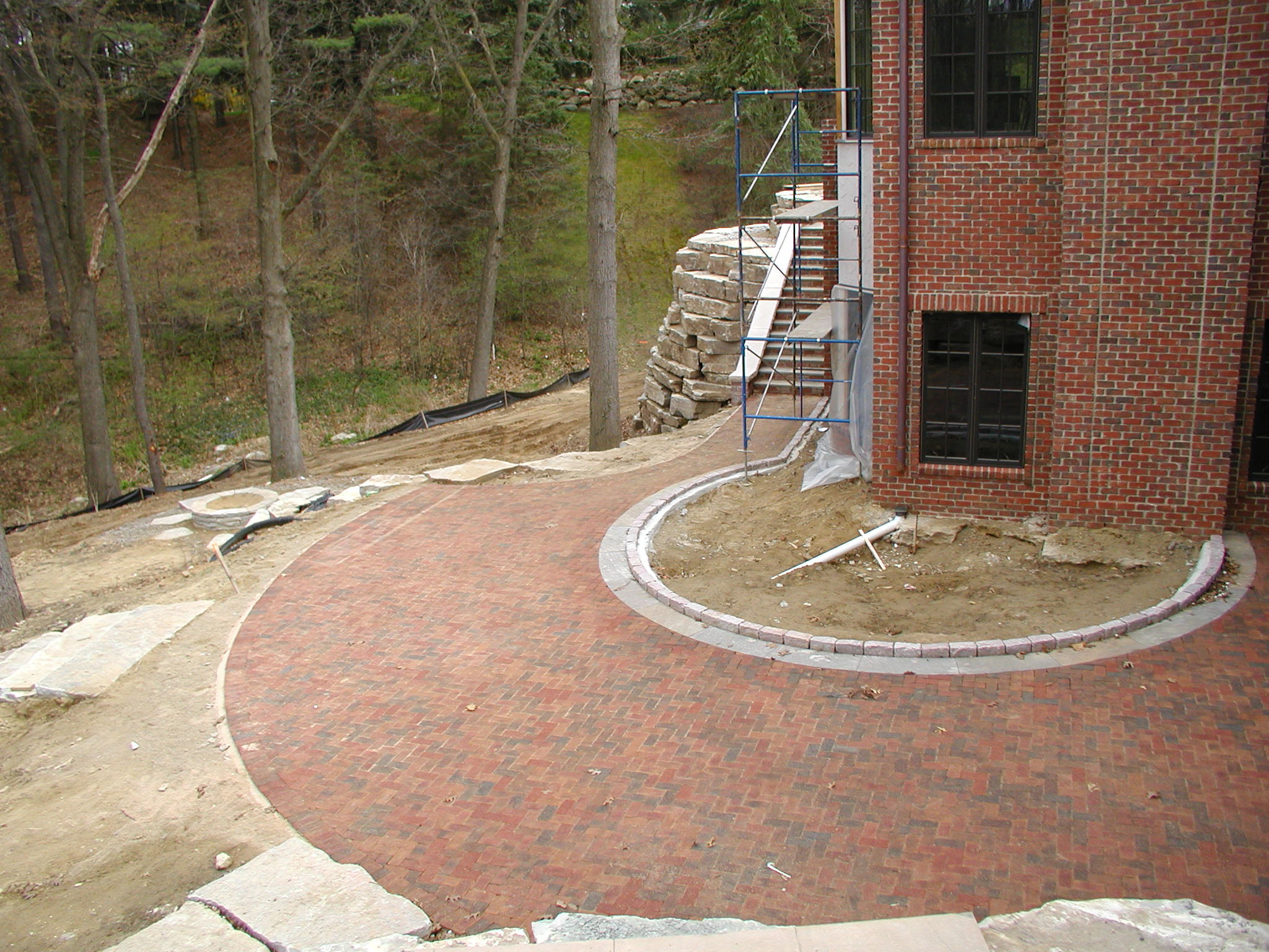 A brick patio is under construction alongside a red brick building, with a scaffold and nearby wooded area visible in the background.