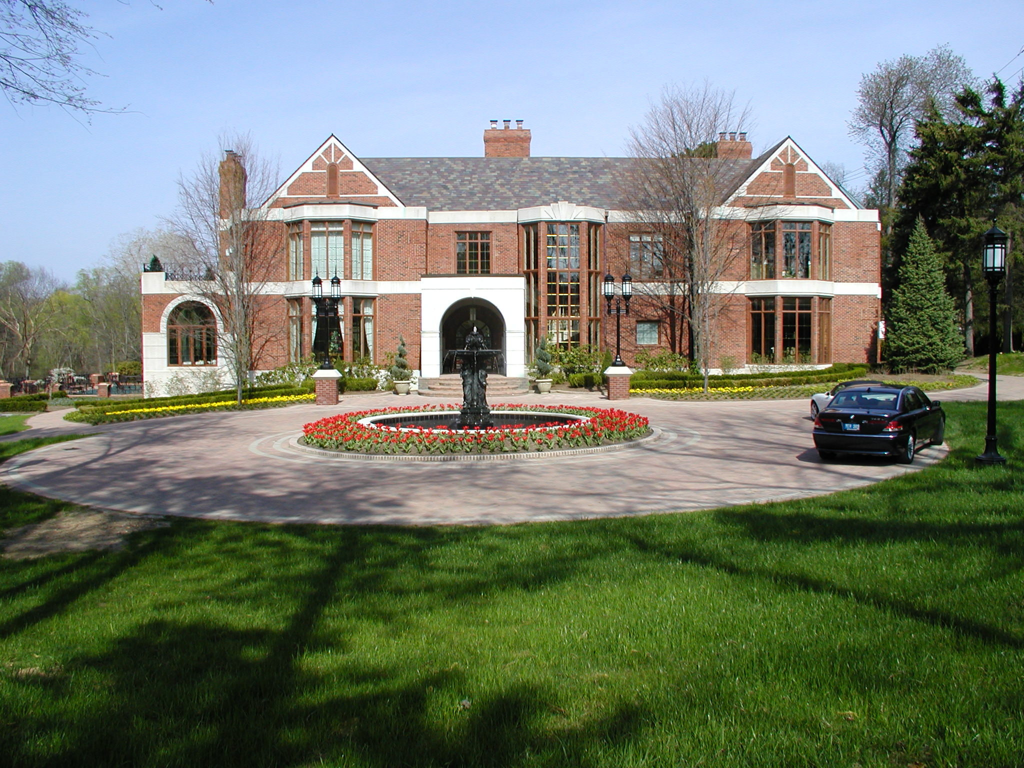 A large brick mansion with extensive windows stands behind a circular driveway with a fountain and colorful flowers, surrounded by trees.