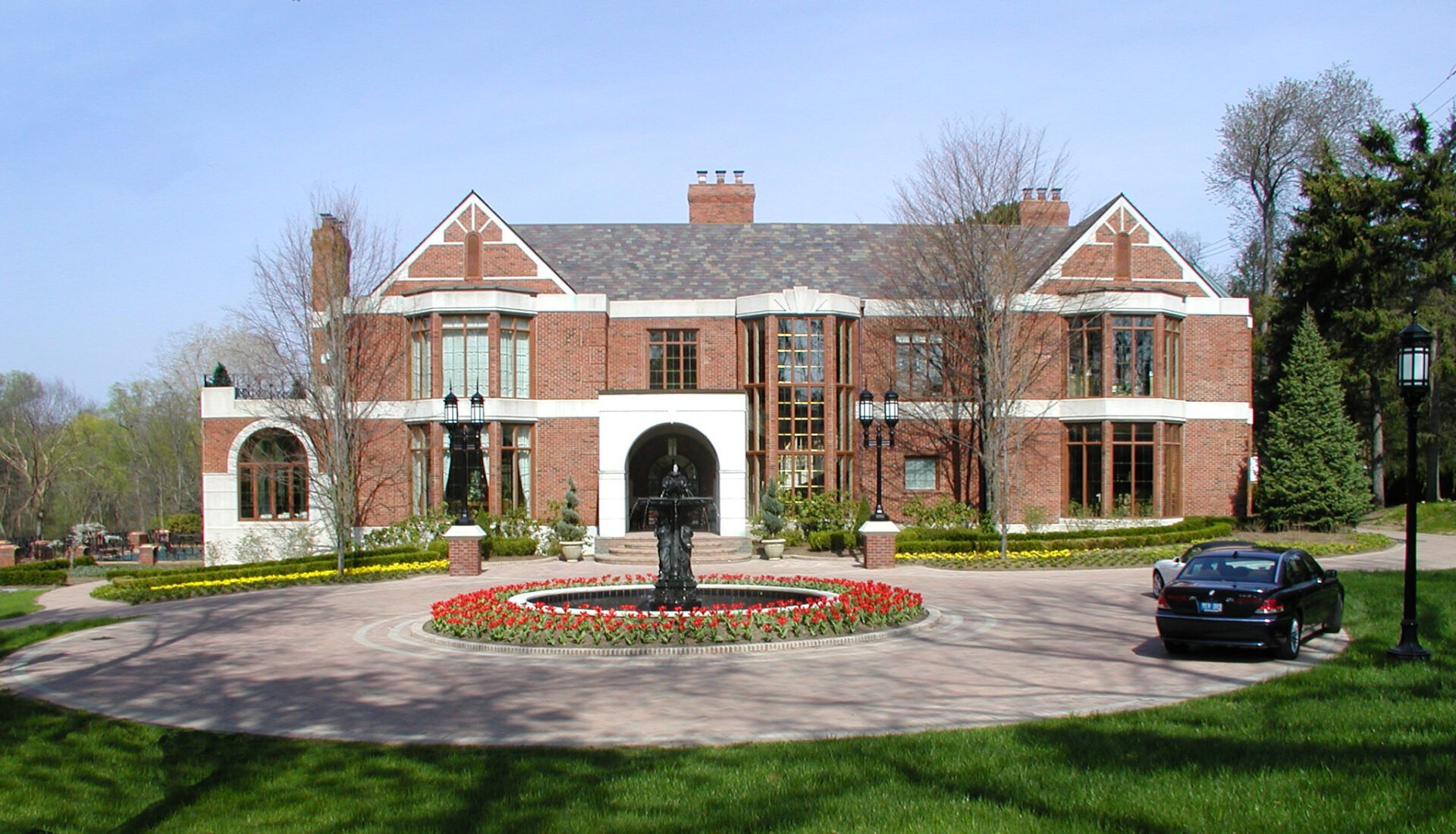 A large red-brick house with a circular driveway featuring a central fountain, surrounded by well-maintained gardens and a parked car.