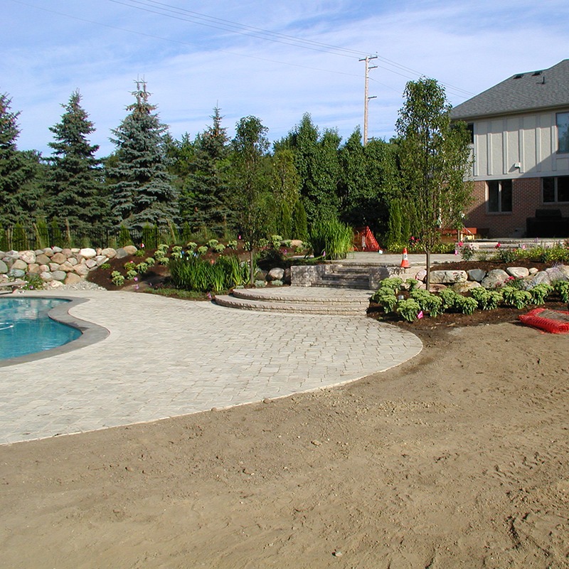 A backyard features a swimming pool, paved patio, lush greenery, and a brick house, with a clear blue sky overhead.