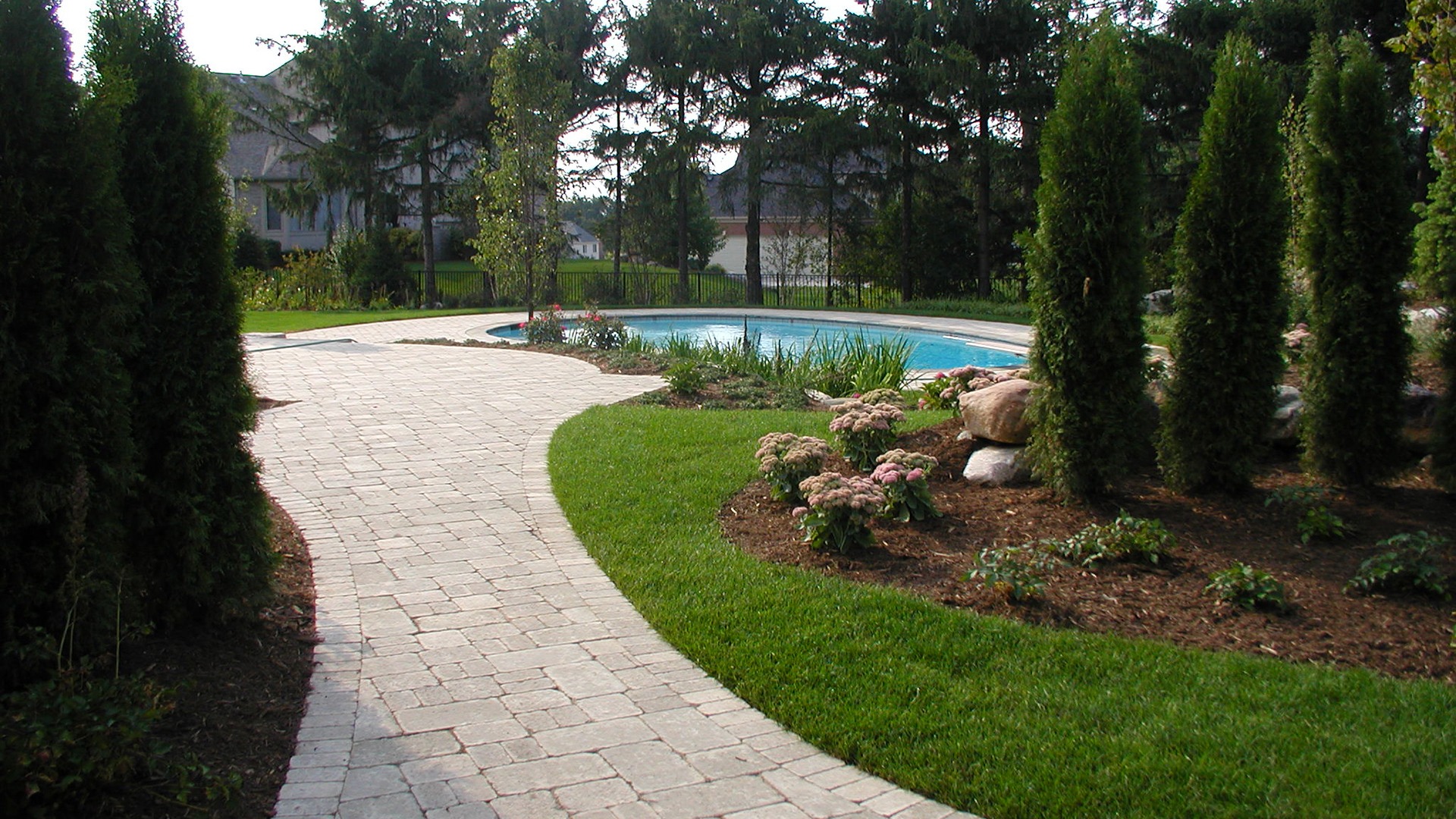 A well-maintained garden features a curved stone pathway leading to a round swimming pool, surrounded by lush greenery and tall trees.
