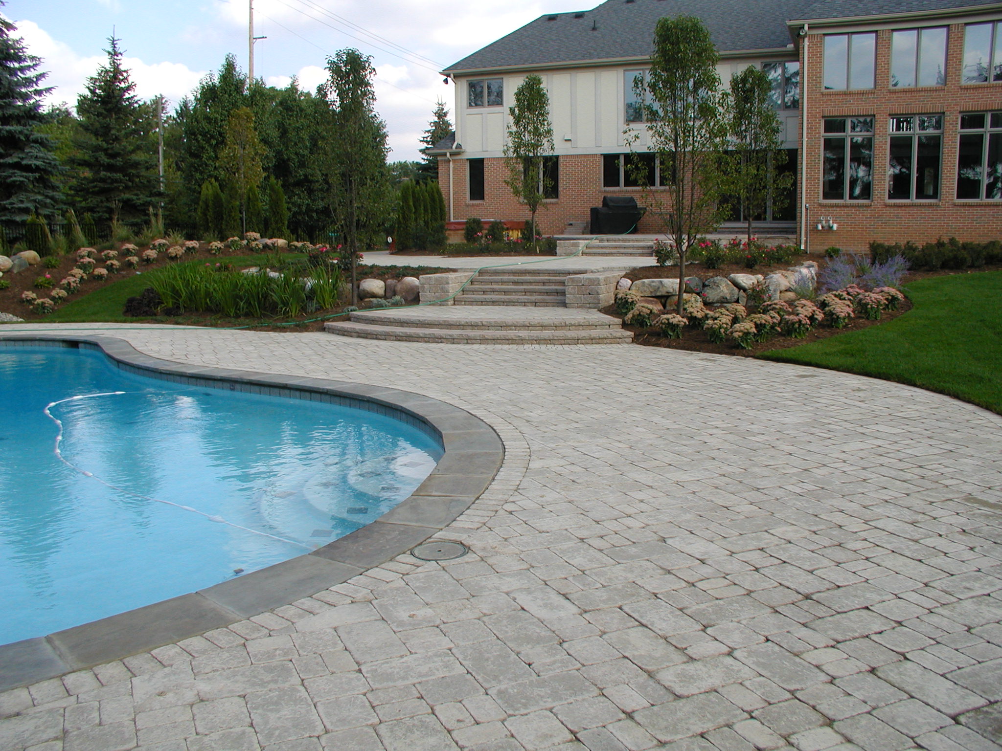 A large brick house features a landscaped backyard with a kidney-shaped swimming pool, stone patio, neat garden beds, and surrounding trees.