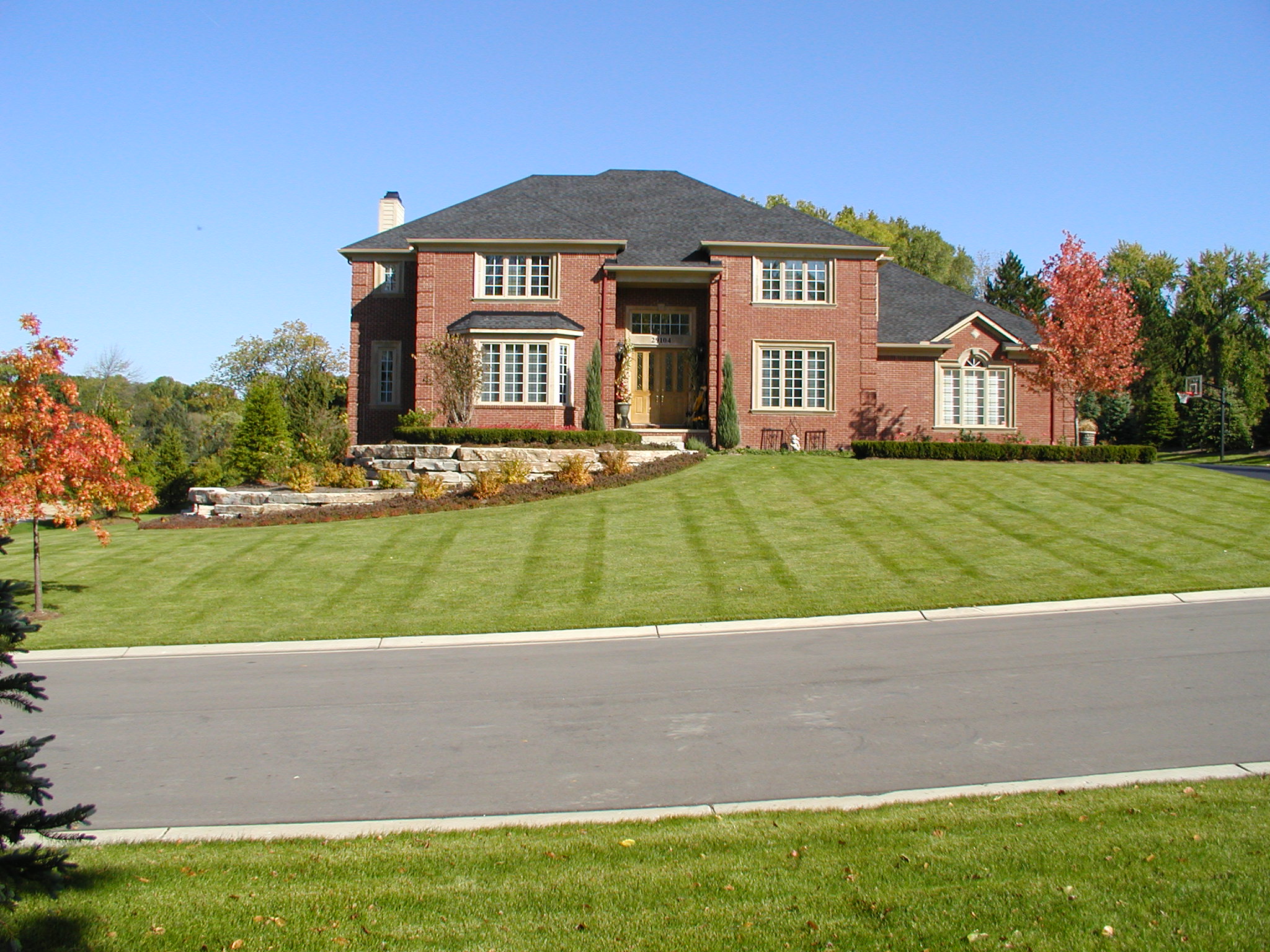 A large, elegant brick house with manicured lawn on a clear, sunny day. Surrounding trees show autumn colors, enhancing the scenic view.