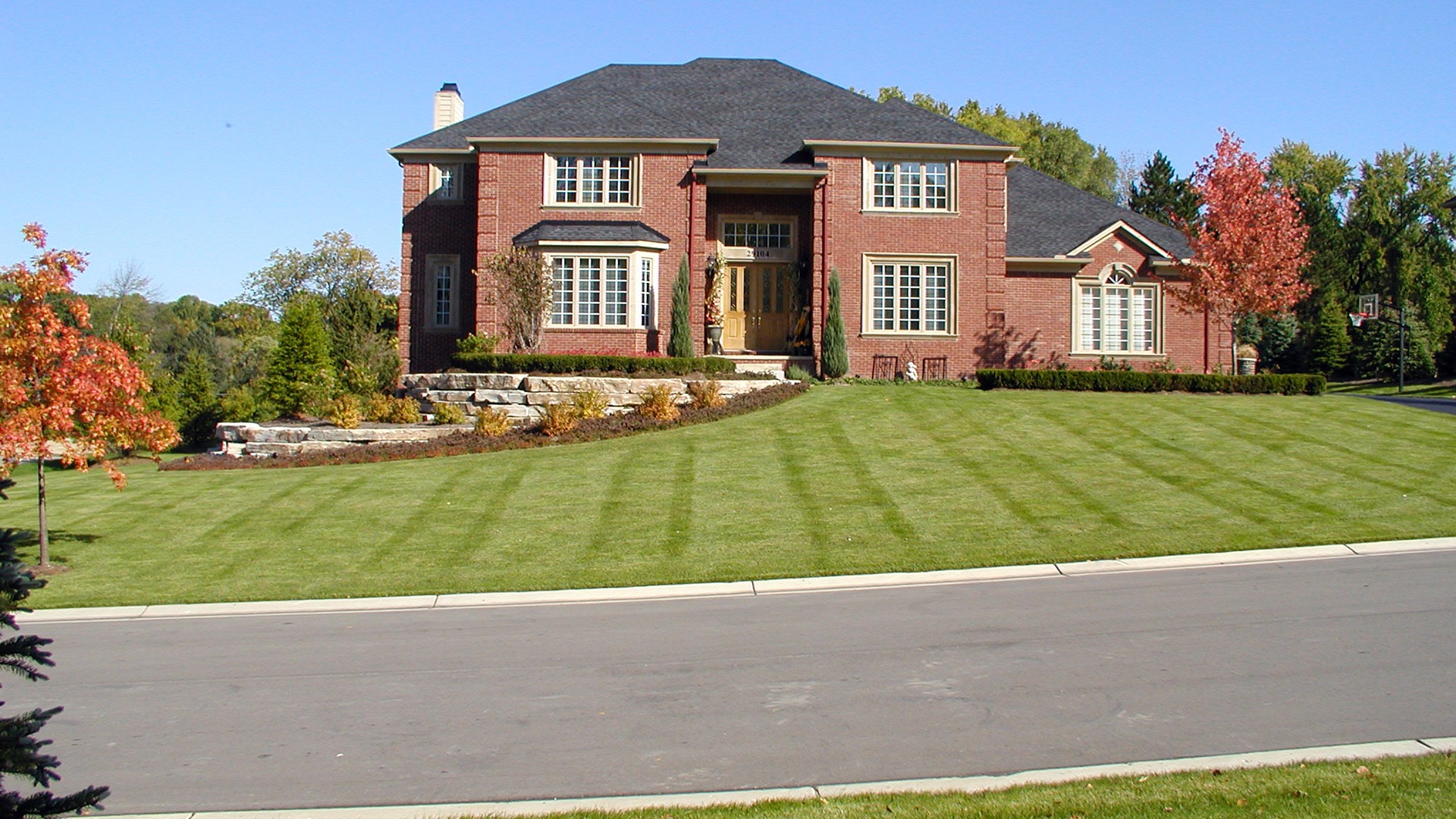 A two-story brick house with manicured lawn, situated on a quiet street. Trees with fall foliage surround the property.