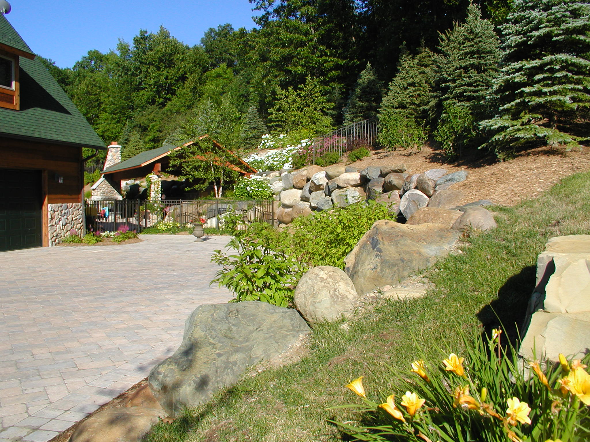 A stone house with a green roof, a paved patio, lush greenery, colorful flowers, and a fenced area adjacent to a wooded hillside.