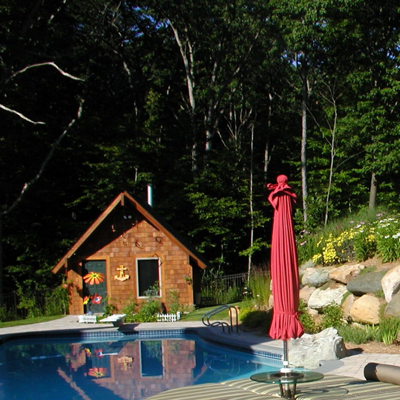 A serene backyard features a pool, red umbrella, and quaint wooden shed surrounded by lush greenery and flowers under clear skies.
