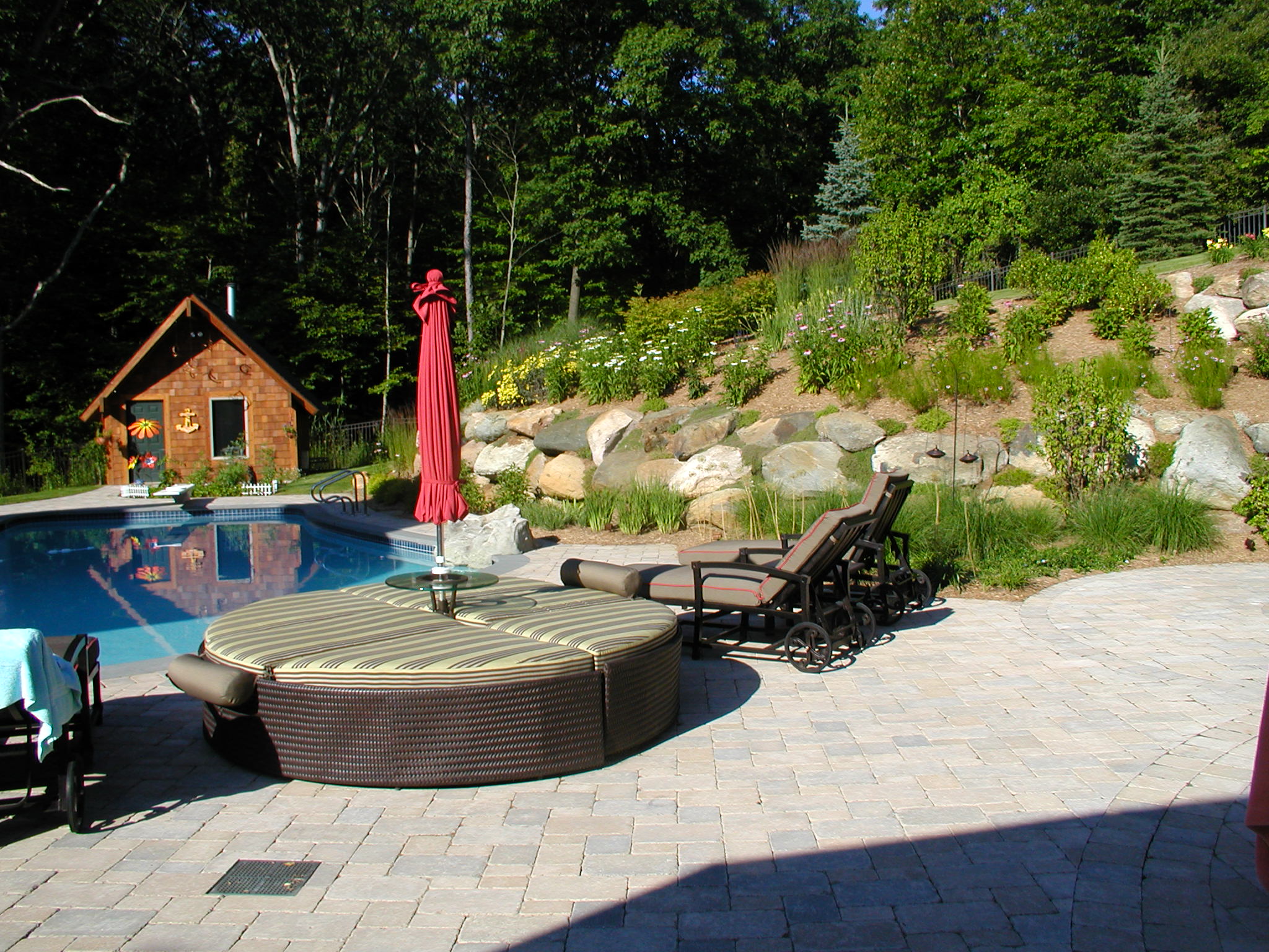 A serene backyard features a swimming pool, lounge chairs, a round daybed, an umbrella, and a quaint cottage surrounded by lush greenery.