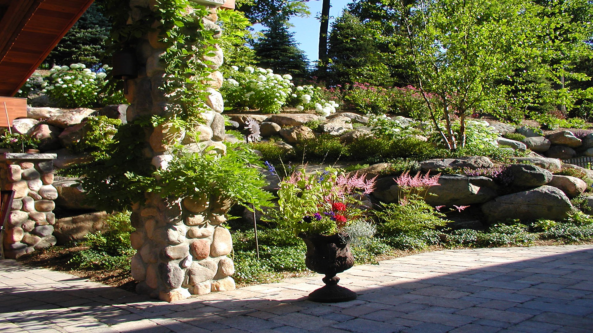 Sunlit garden with colorful flowers, stone pillars, and greenery surrounding a paved patio area, providing a peaceful, natural setting under clear skies.