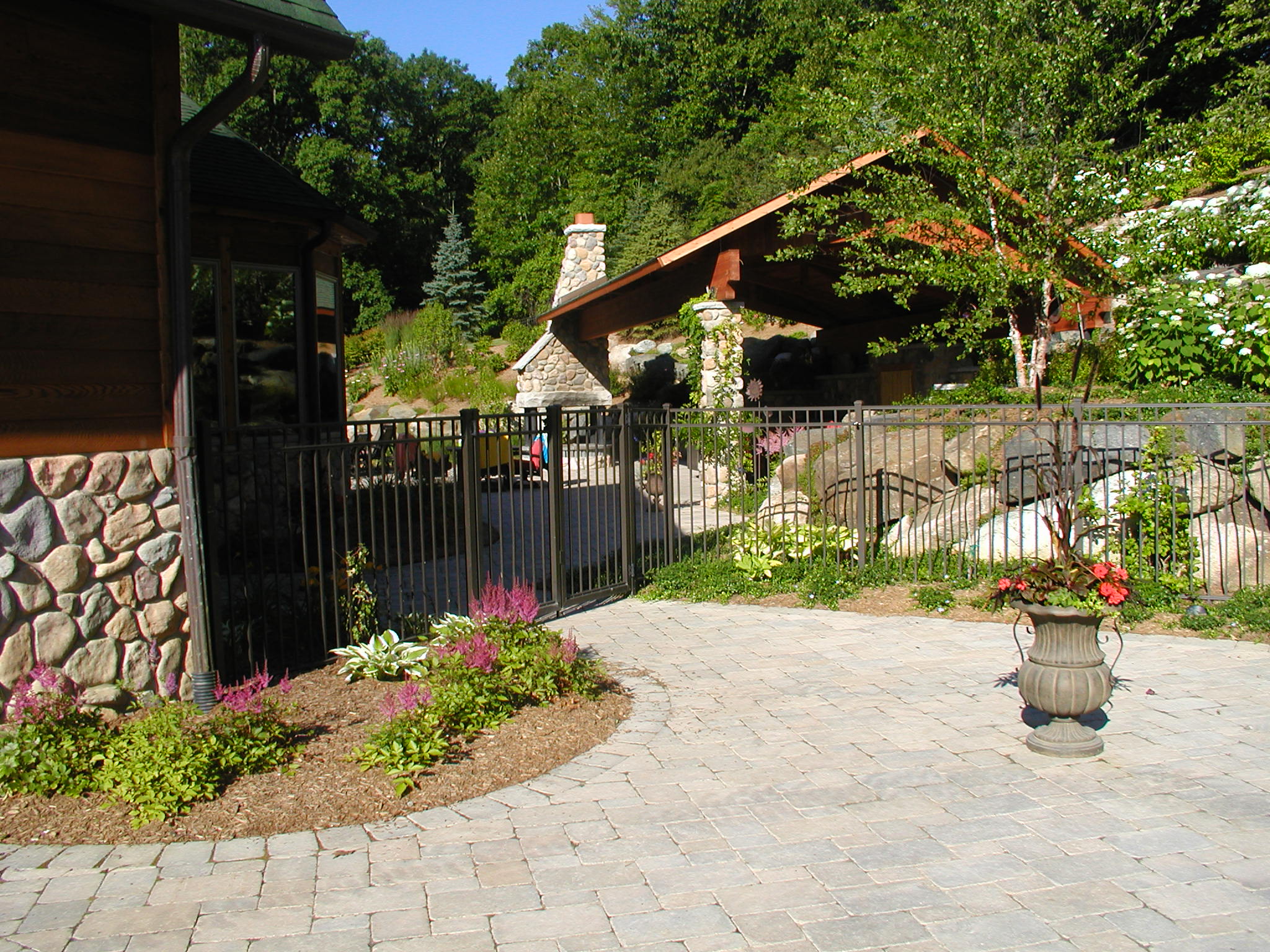 A serene garden area with stone buildings and lush greenery, featuring colorful flowers, a paved walkway, and a fenced enclosure.