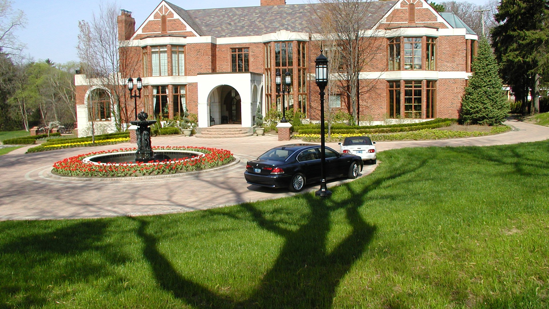 A large brick mansion with a central fountain, two parked cars, and well-maintained gardens in a lush, green setting.