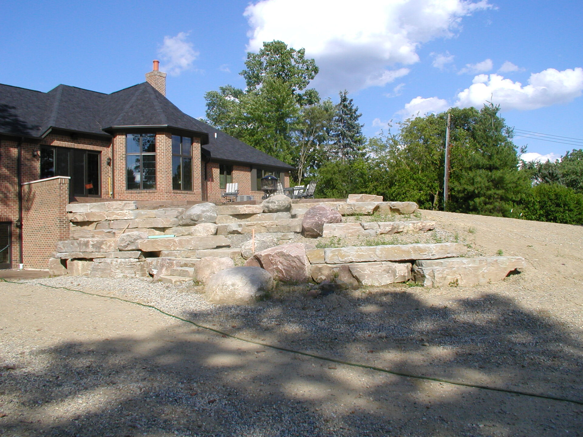 Brick house with large windows overlooks a tiered stone patio. Surrounded by trees, the landscape includes gravel and a blue sky with clouds.