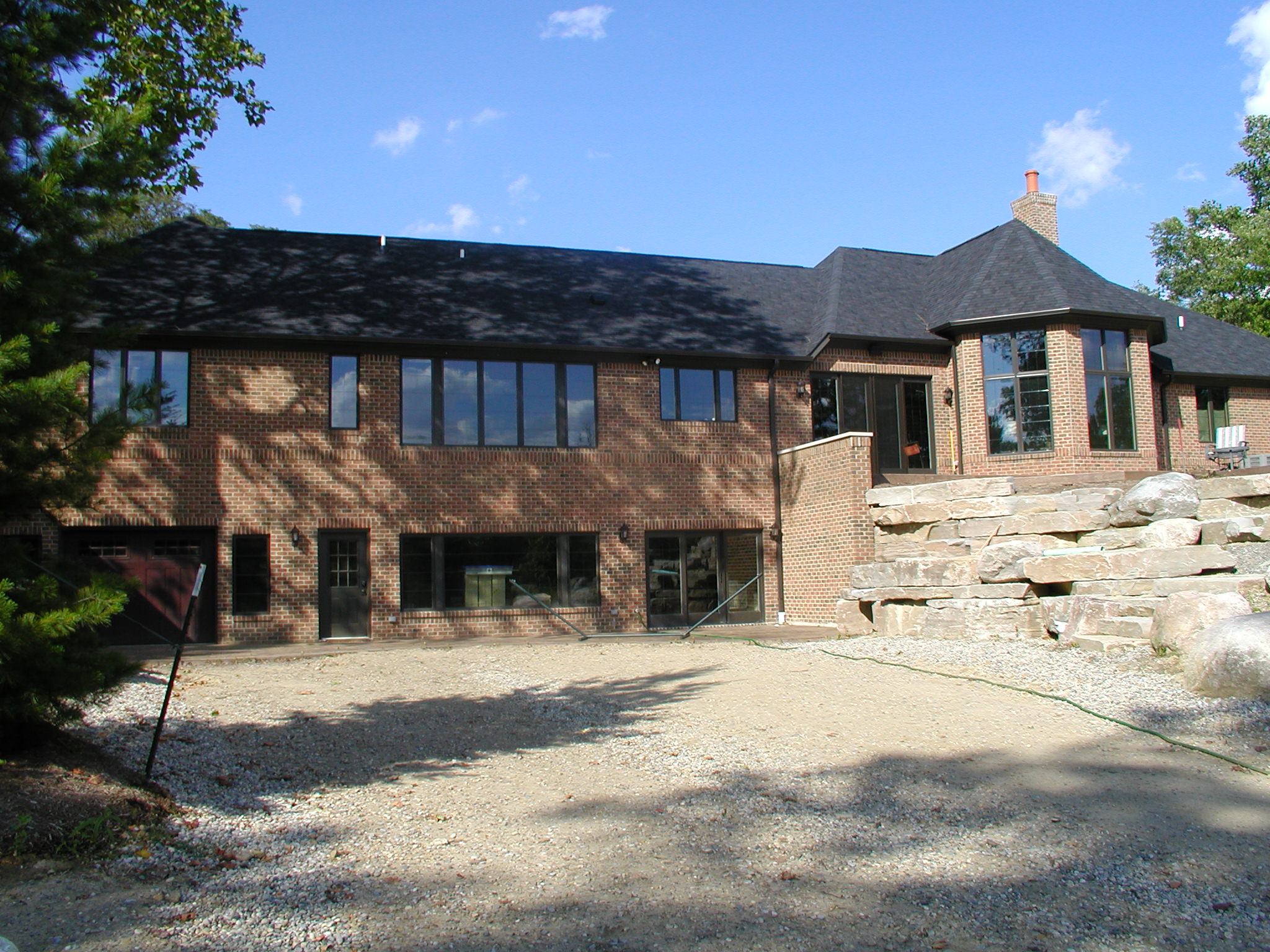 A large brick house with numerous windows, surrounded by stone landscaping and trees, under a clear blue sky with scattered clouds.