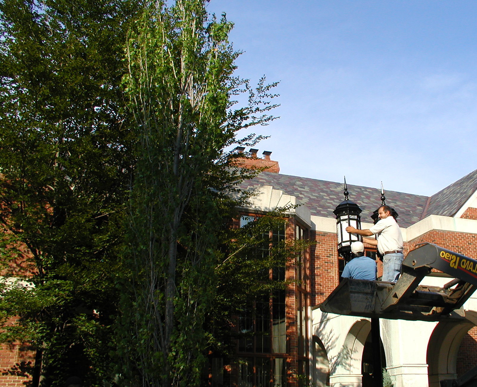 Two people work on a lamppost using a lift near a brick building, surrounded by trees. New Holland machine visible.