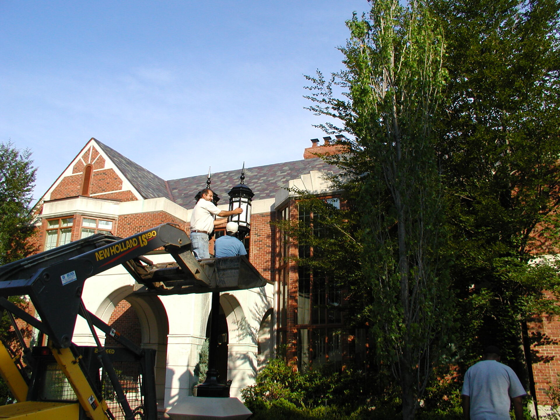 Two people on a lift work on a lantern outside a brick building, surrounded by trees, under a clear blue sky.