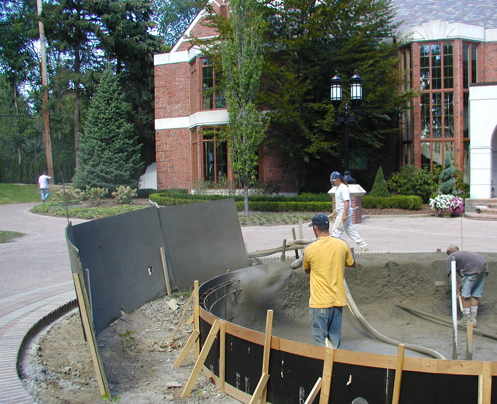 Three people work on concrete construction in front of a brick building with large windows and landscaped surroundings.