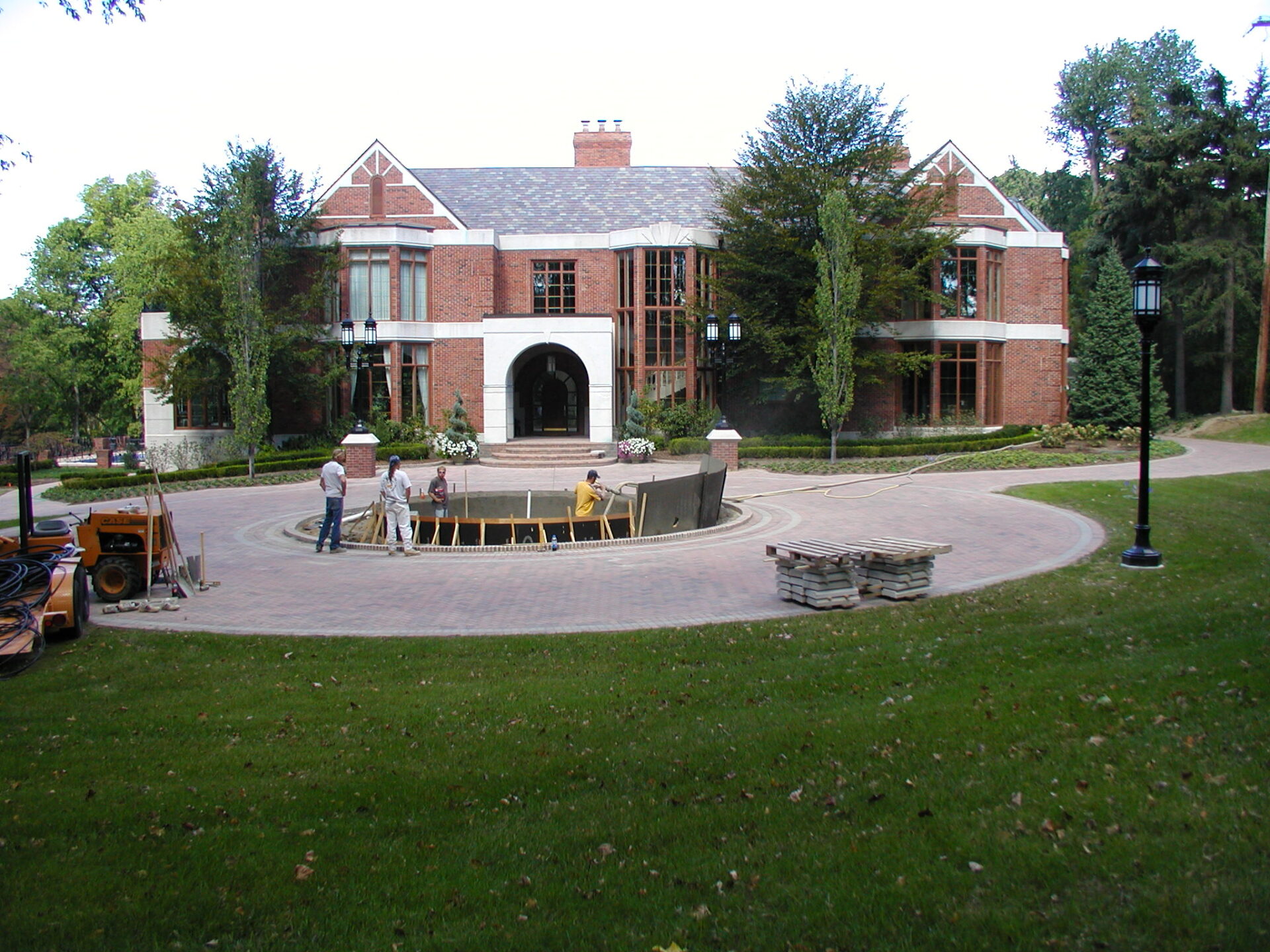 A group of people works on a construction project in front of a large, brick building surrounded by trees and a well-maintained garden.