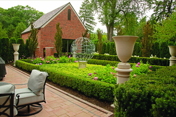 A beautiful garden features manicured hedges, a geometric sculpture, brick building, and patio chairs, creating a serene and picturesque outdoor space.