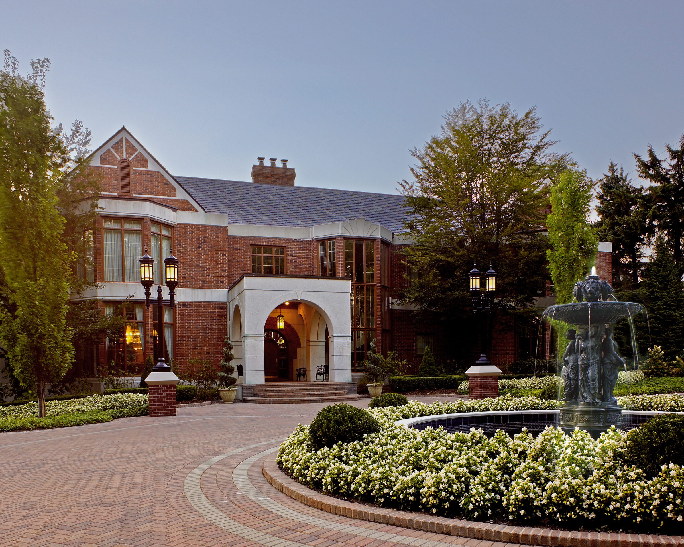 Brick building with arched entrance, surrounded by lush greenery and ornate fountain in foreground. Elegant street lamps illuminate the scene.
