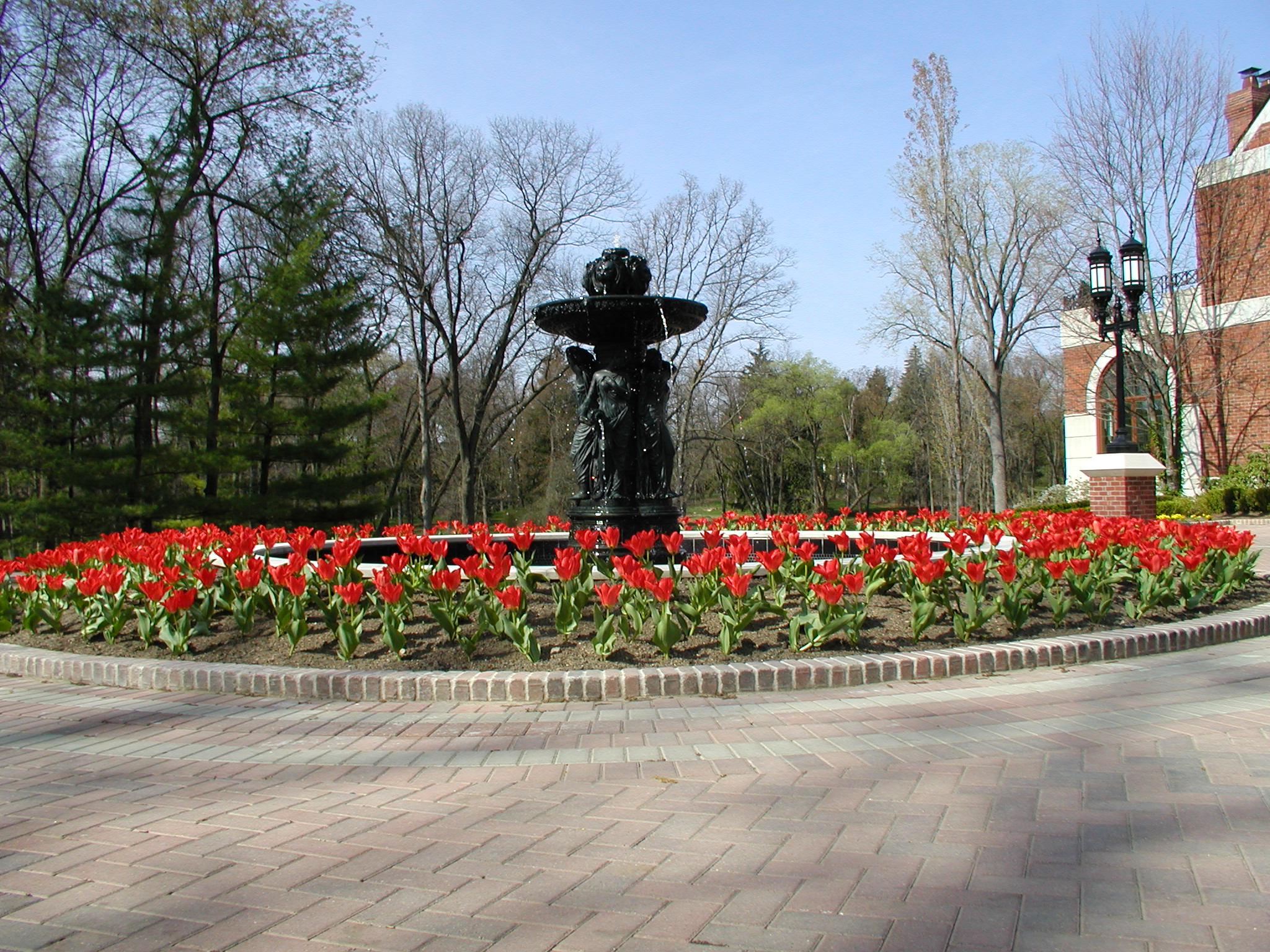 A black fountain surrounded by red tulips is in front of a brick building, amidst tall trees and clear blue sky.