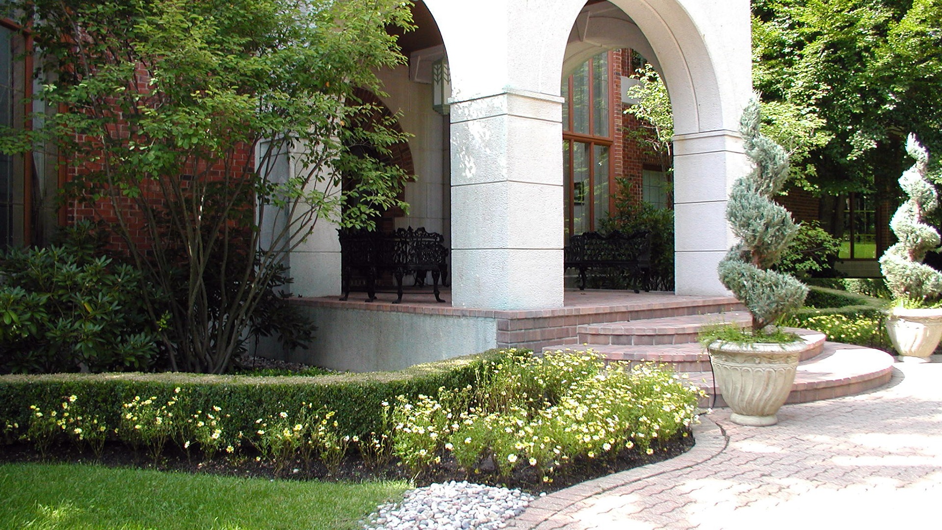 The image shows a garden entrance with arches, brick steps, decorative plants, and wrought-iron benches, surrounded by lush greenery and manicured bushes.