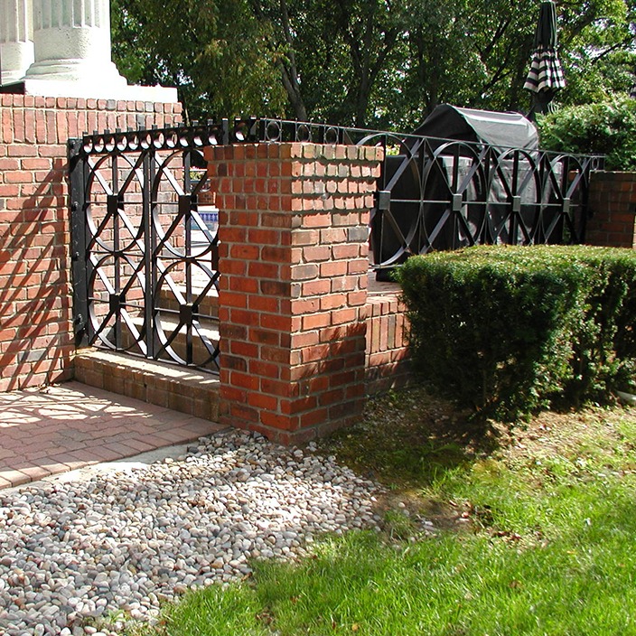 A brick pillar with iron railing leads to a raised patio. Lush greenery and small pebbles adorn the landscape around it.