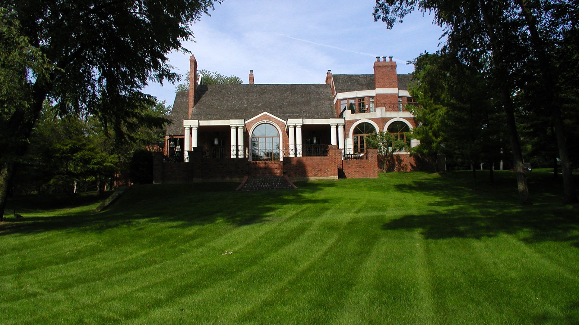 Historic brick mansion with large windows and columns set in a lush green landscape, surrounded by tall trees under a clear blue sky.