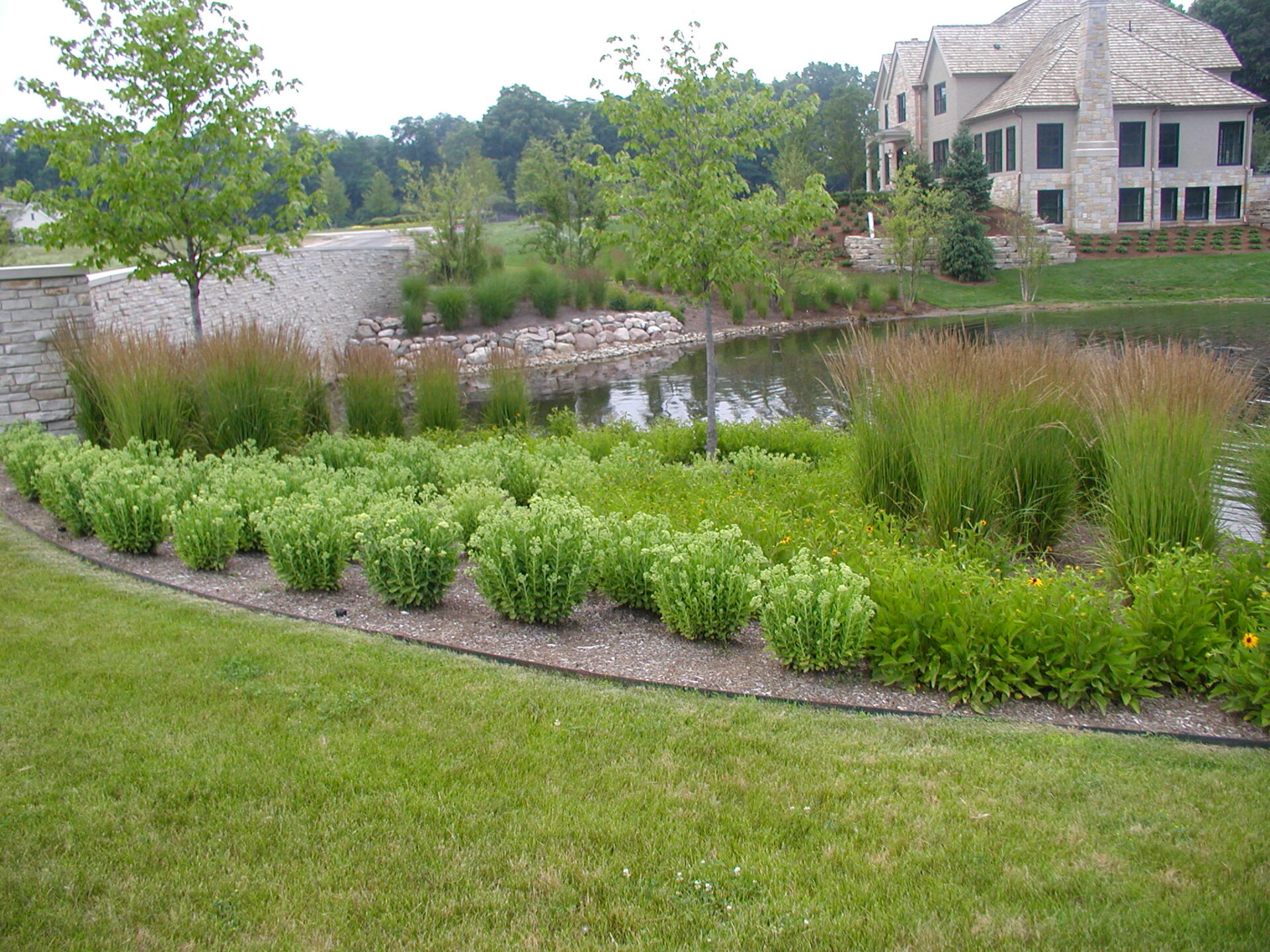 A landscaped garden with lush greenery surrounds a stone building, overlooking a serene pond and lined with small trees and shrubs.