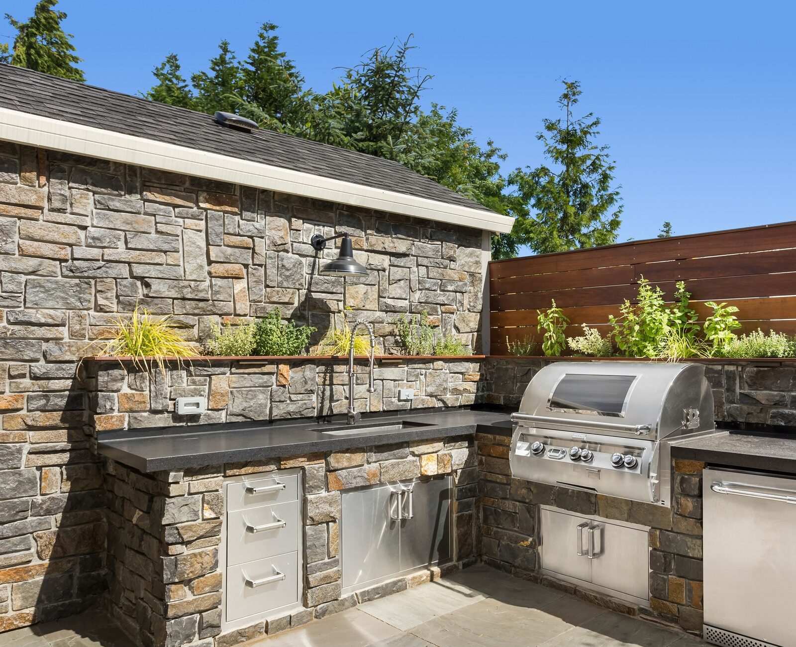 Outdoor kitchen features stone counters, stainless grill, and cabinets, surrounded by plants and wooden fence against a backdrop of trees and clear sky.