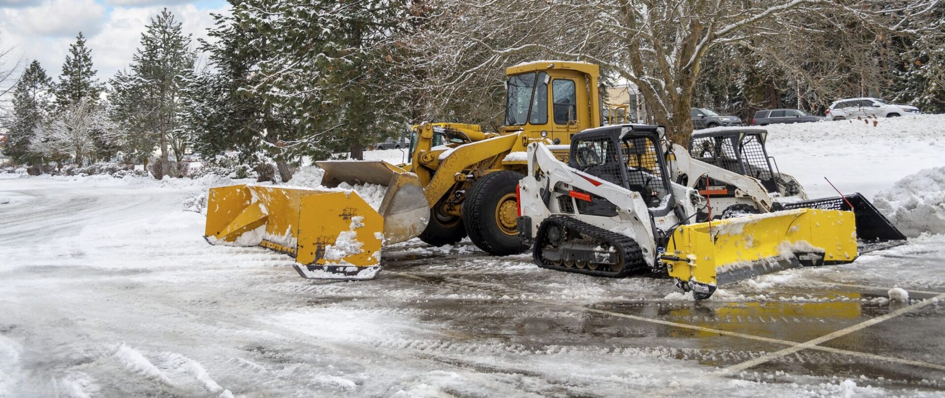 Snow-covered parking lot with two snow plows and a loader. Trees and parked cars in the background, creating a snowy winter scene.