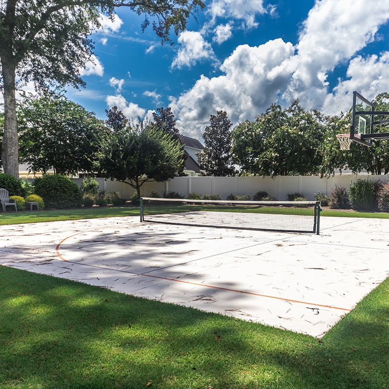 A sunny backyard features a multi-use sports court with a basketball hoop and net, surrounded by trees and a white fence.