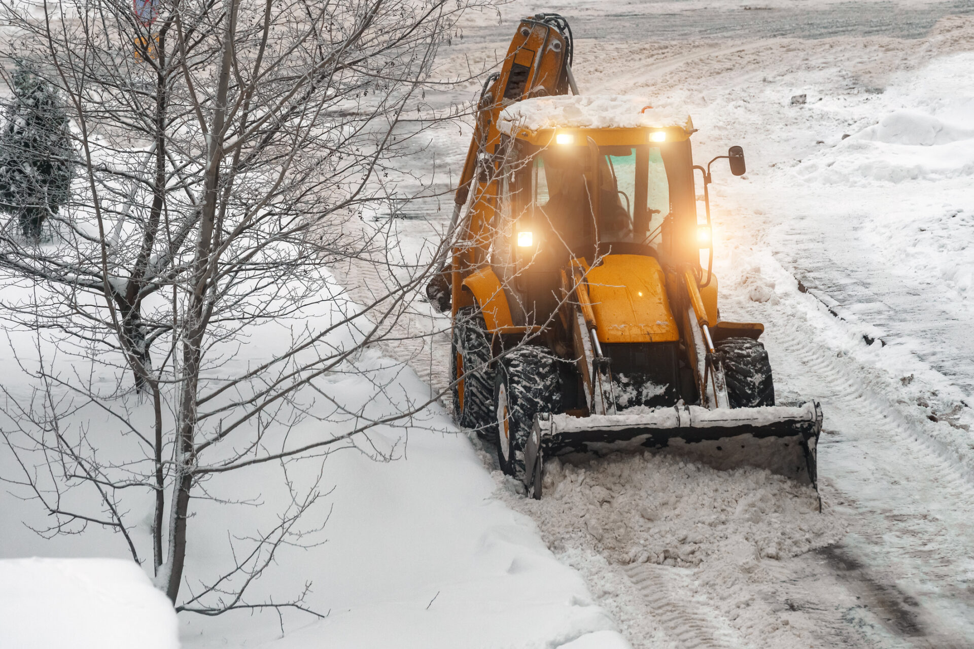 Yellow snowplow clearing a snowy road, surrounded by snow-covered trees and pavement. The vehicle's lights are on, illuminating its path.