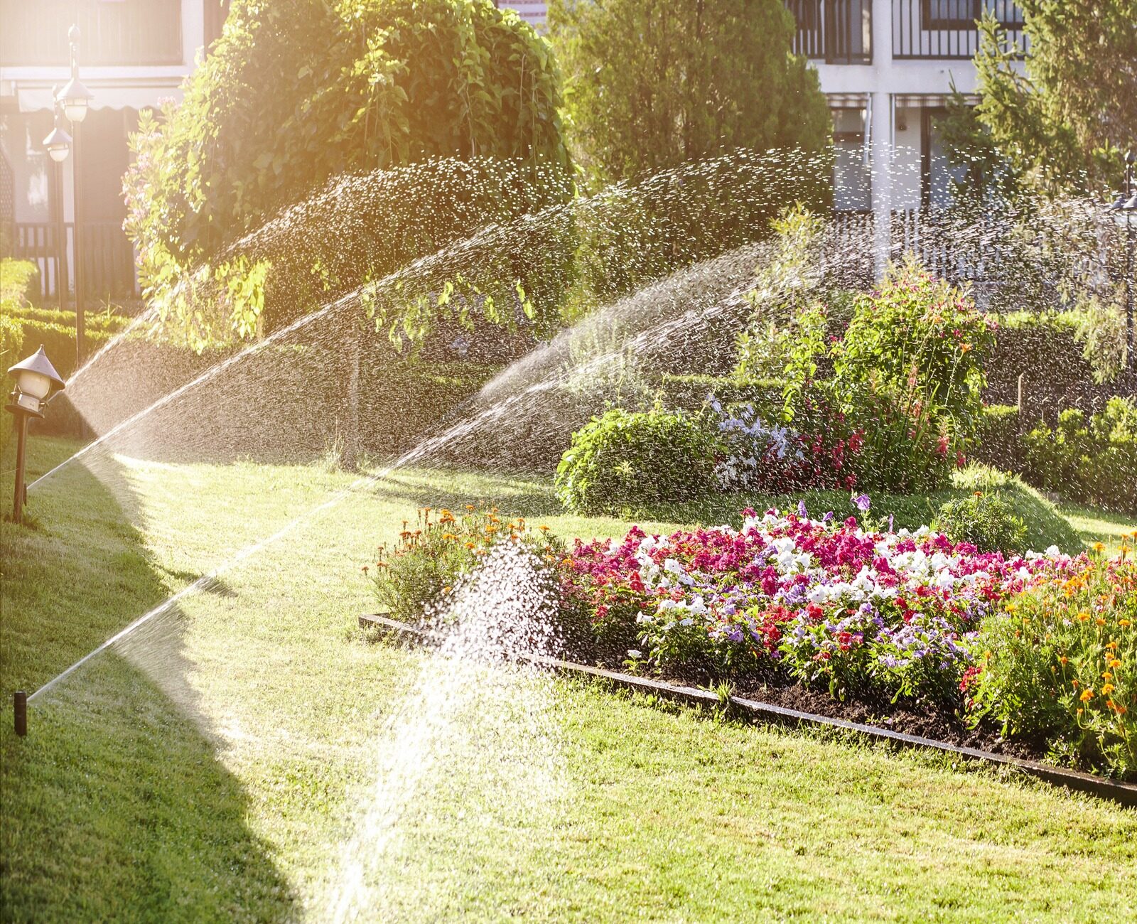 Sprinklers water vibrant flowerbeds in a sunlit garden, with lush greenery and a building in the background, creating a serene atmosphere.