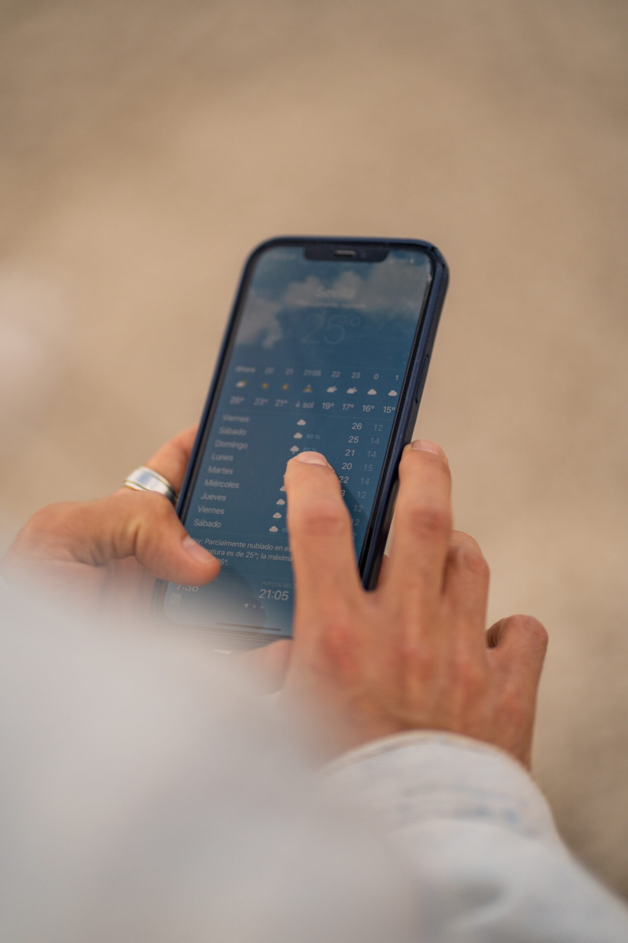 A person uses a smartphone to check the weather forecast for Seville, showing temperature and weekly weather icons on the screen.