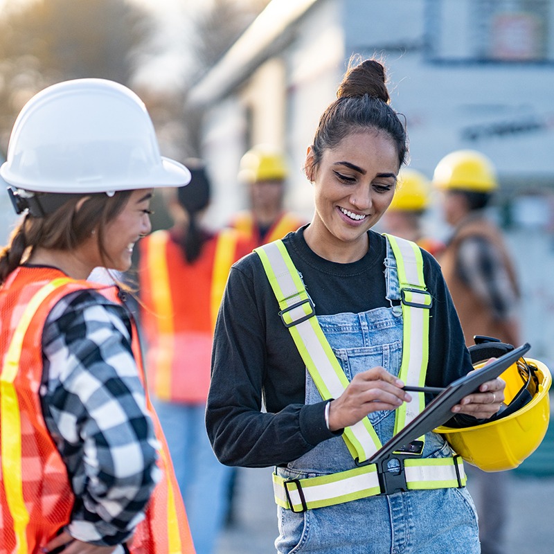 Two people wearing safety gear and vests, smiling, in a construction setting. One holds a tablet; hard hats are visible.