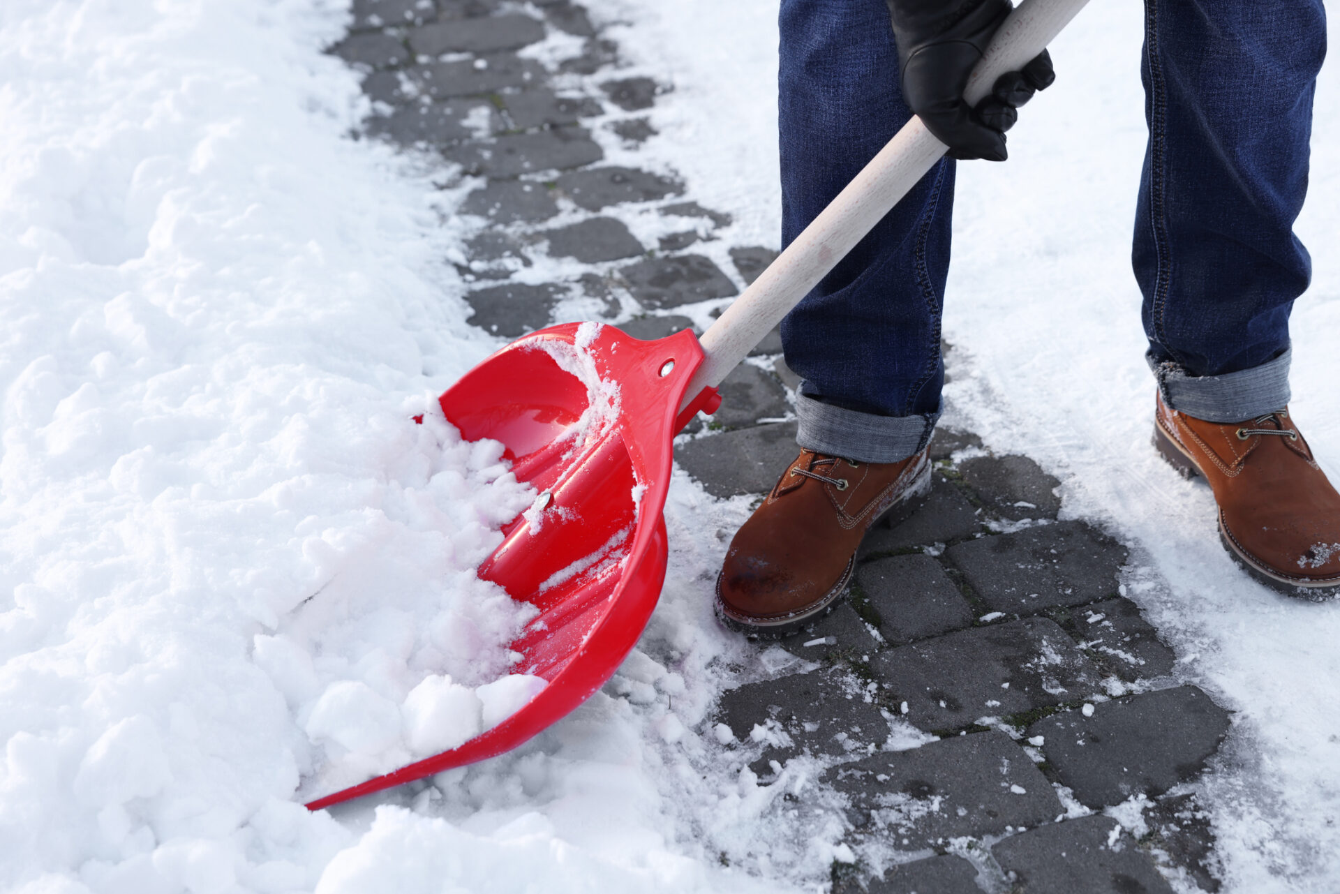 A person wearing brown boots shovels snow with a red shovel on a stone path, showing a winter scene in progress.