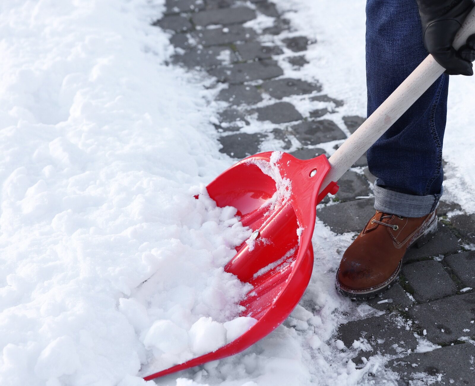A person wearing brown boots shovels snow with a red shovel on a stone path, showing a winter scene in progress.