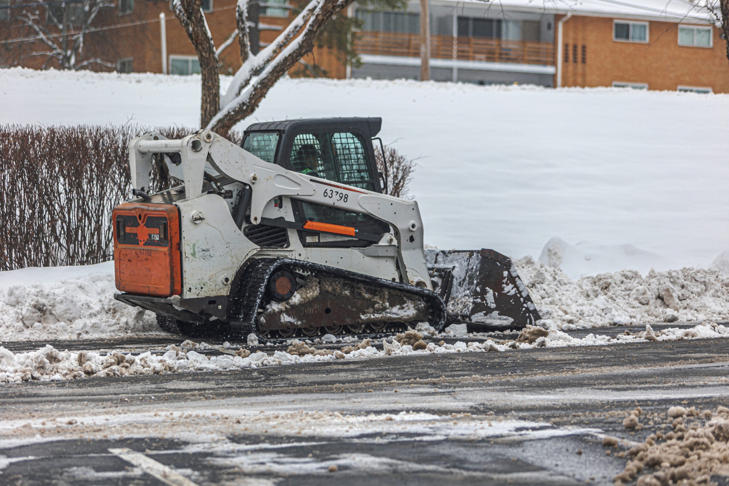 A skid steer loader clearing snow from a residential street with snow-covered houses in the background, under overcast sky conditions.