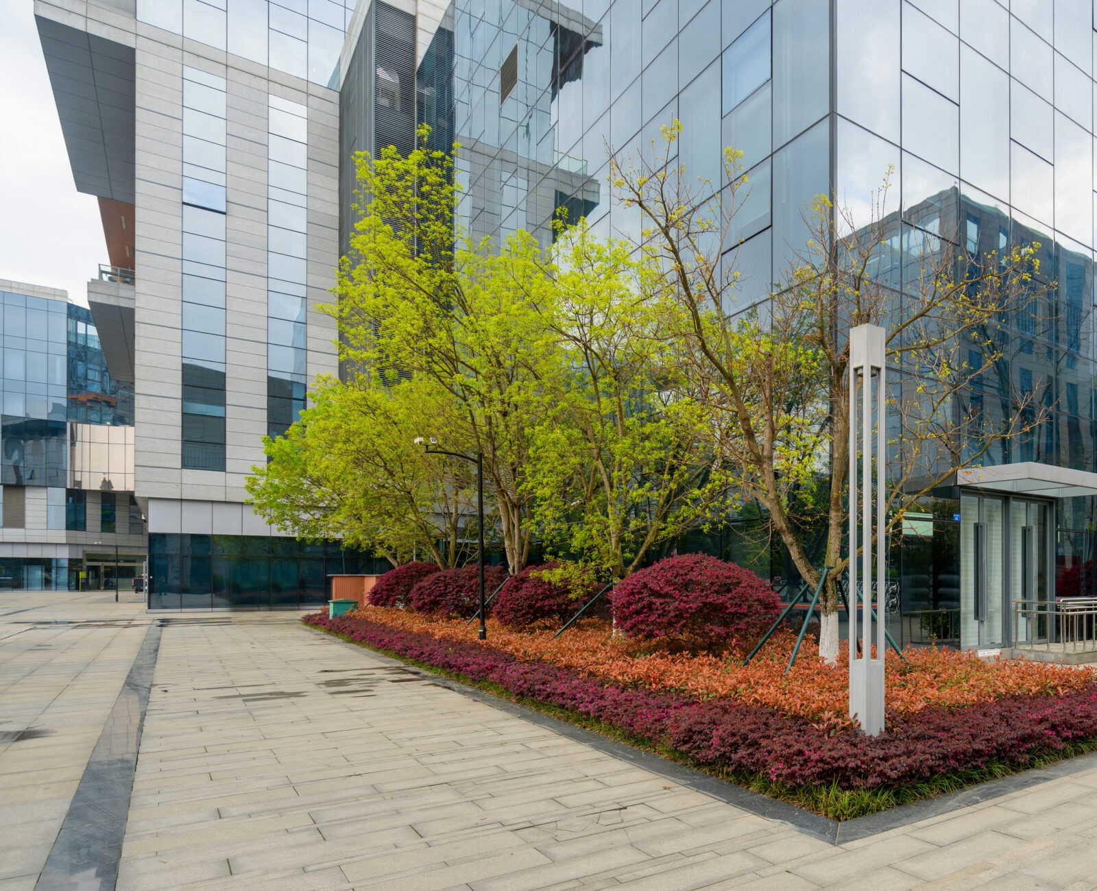 Modern glass building with landscaped garden, featuring green and reddish foliage, paved walkway, and surrounding skyscrapers. Urban architecture and natural elements harmonize.