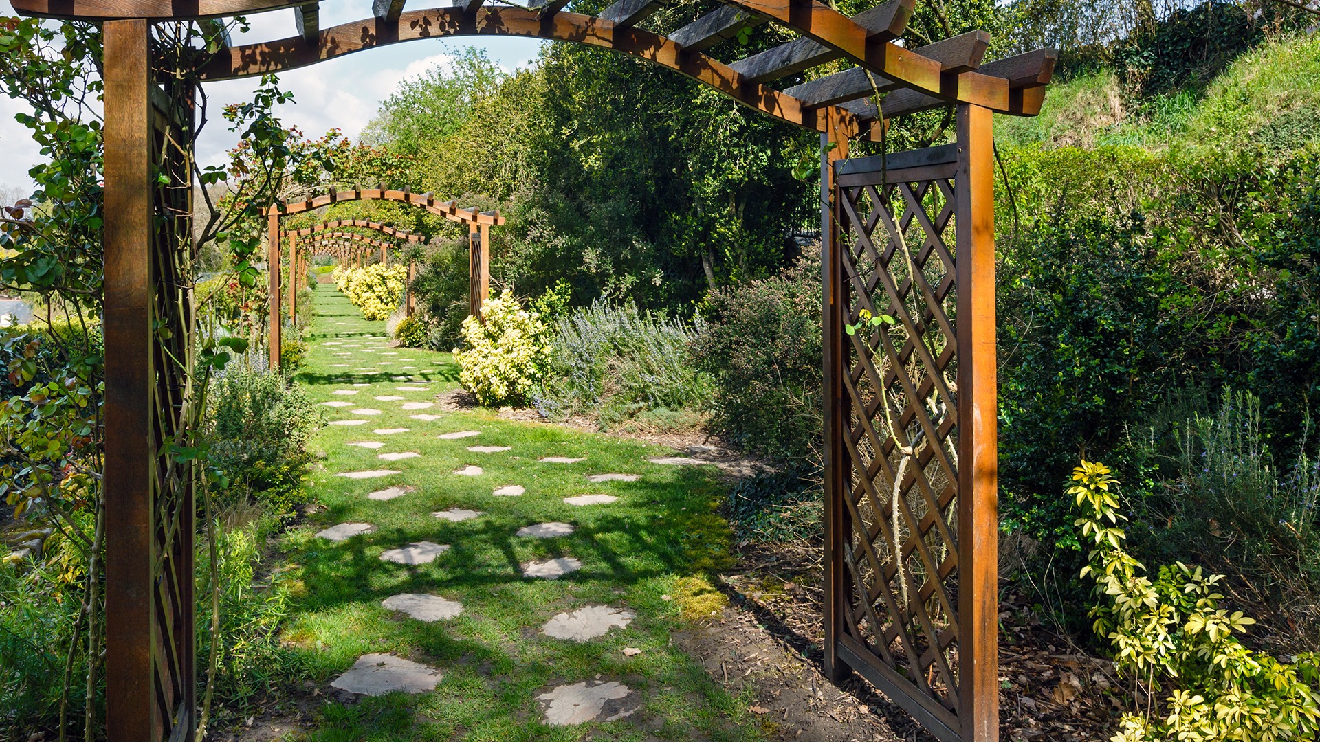 A serene garden with wooden archways and stone pathway, surrounded by lush greenery and vibrant shrubs beneath a clear blue sky.
