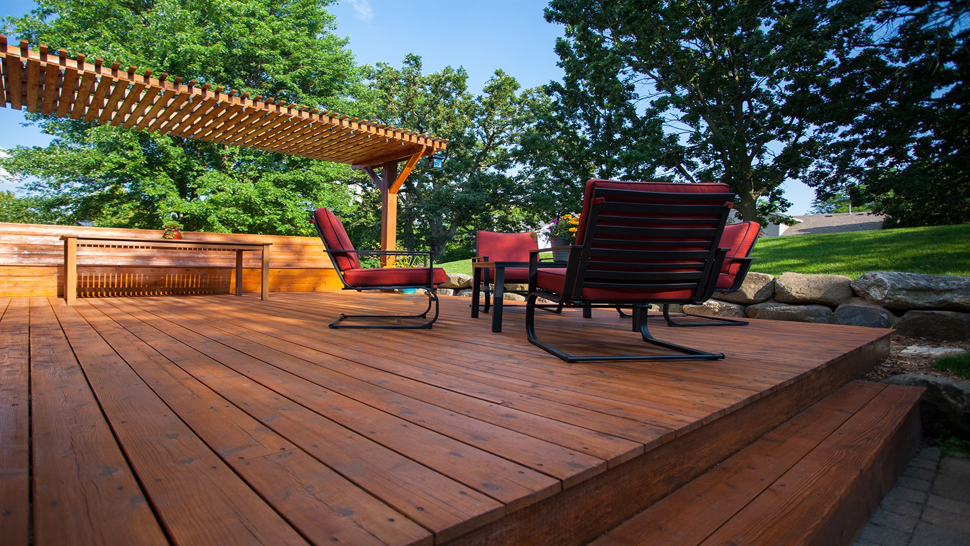 A wooden deck features red cushioned chairs and a table under a pergola, surrounded by lush green trees and stone landscaping.