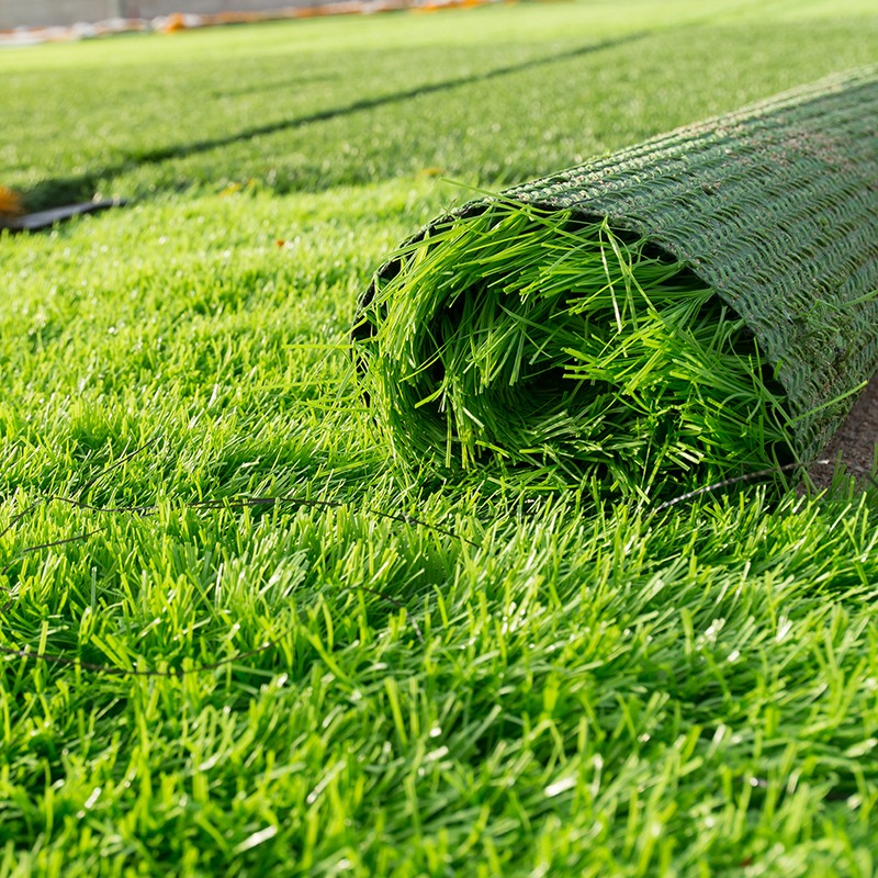 A rolled-up section of artificial green turf lies on a field, highlighting vibrant, closely-packed blades of grass under sunlight.