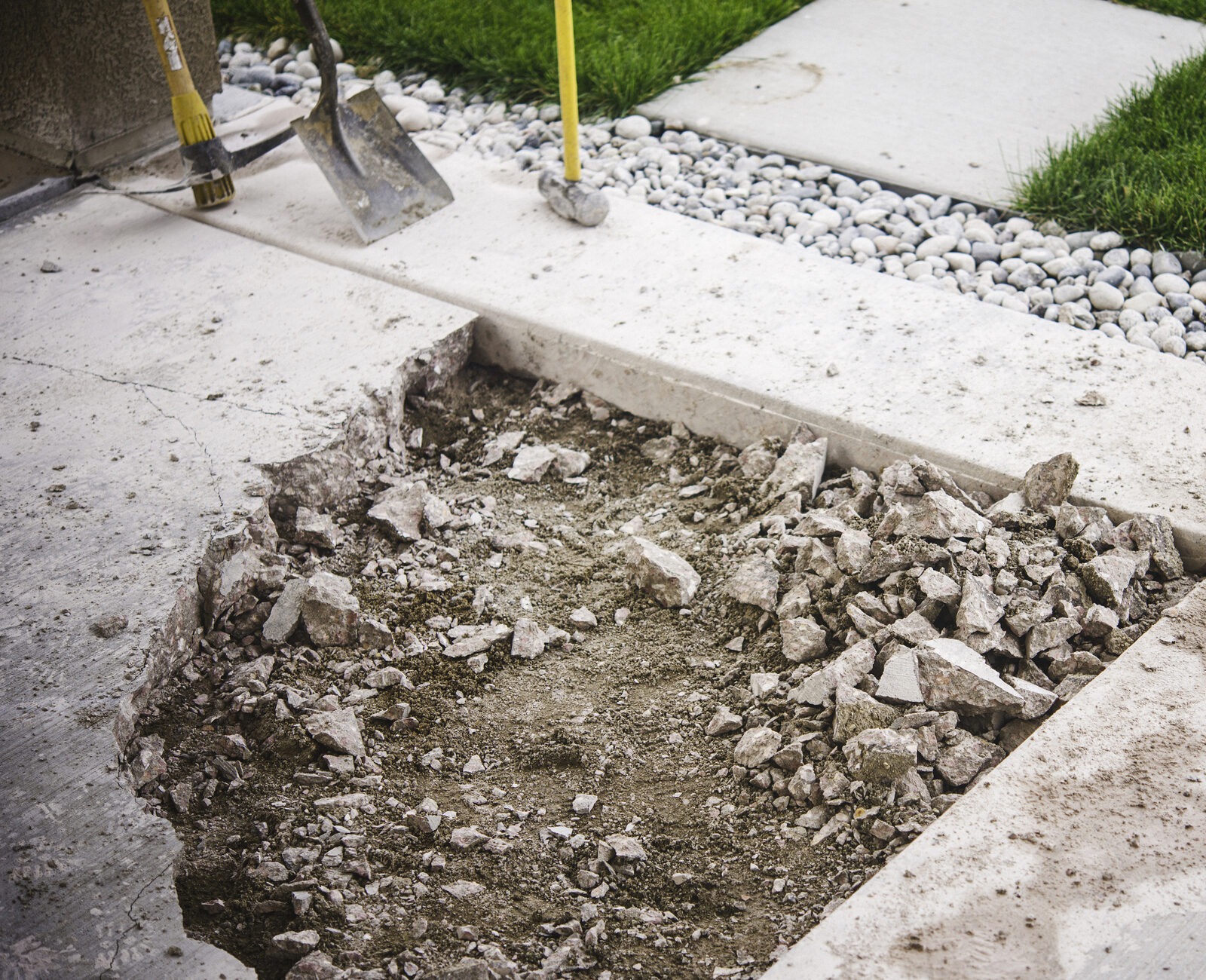 A concrete slab is broken with rubble, surrounded by grass and pebbles. Shovel and sledgehammer visible nearby.