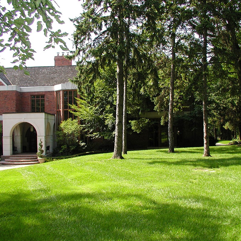 Brick building surrounded by tall trees and lush green lawn, featuring arched entrance and windows allowing natural light. Peaceful and inviting outdoor scene.