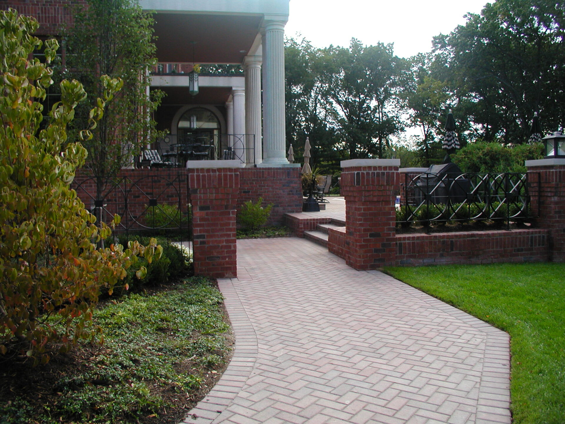 A brick pathway leads to a building with columns and outdoor seating, surrounded by greenery and trees under a clear sky.