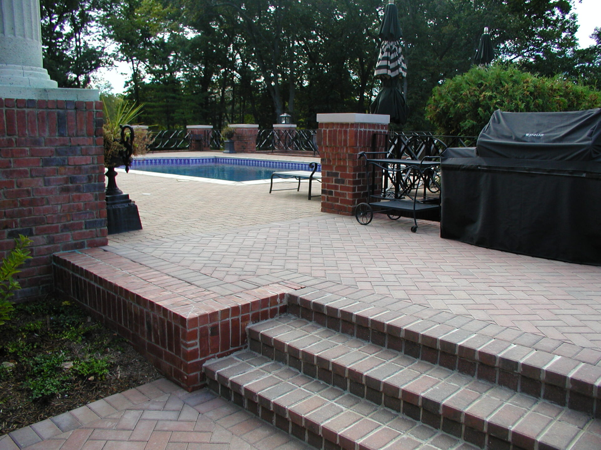 Brick patio with steps, outdoor pool, barbecue grill, and black covered umbrellas. Surrounded by trees, creating a peaceful backyard atmosphere.