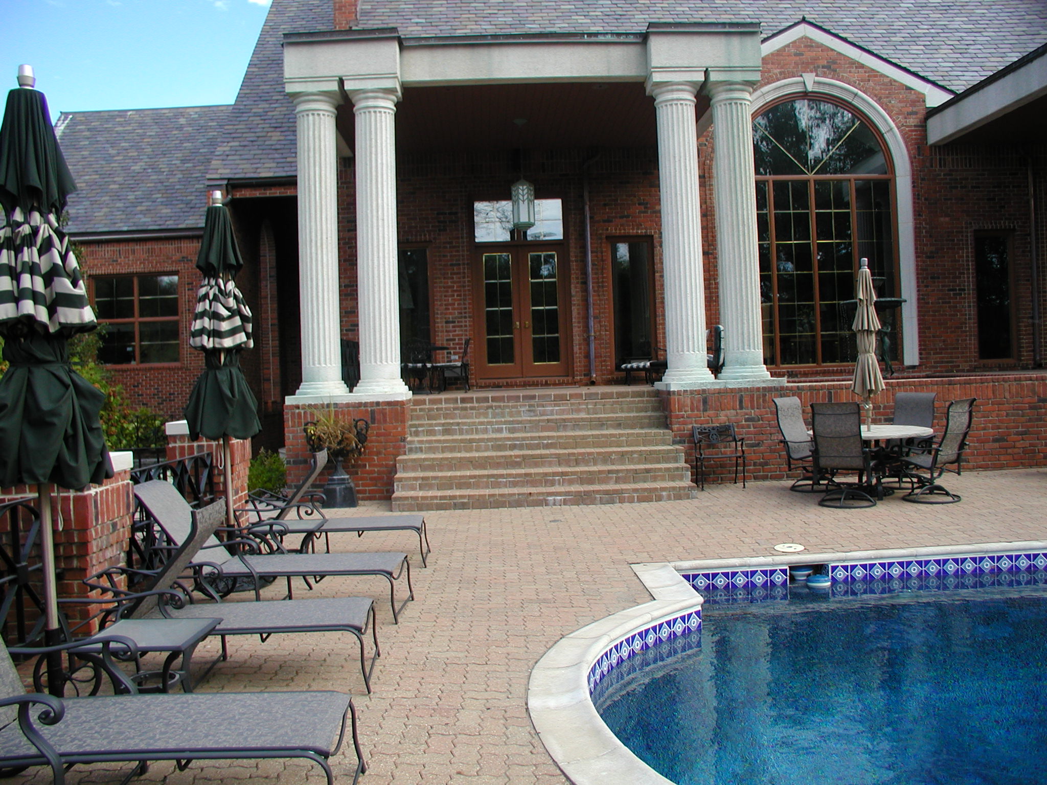 A red-brick house with columns overlooks a pool surrounded by lounge chairs and patio furniture, under cloudy skies.