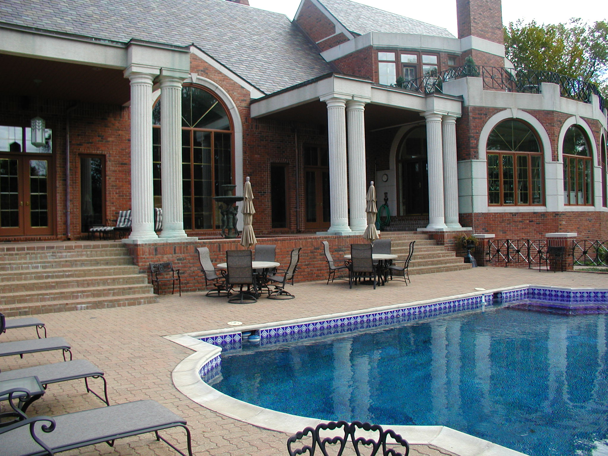 Luxurious brick mansion with large windows, tall columns, and a spacious patio featuring an elegant swimming pool surrounded by lounge chairs and dining tables.
