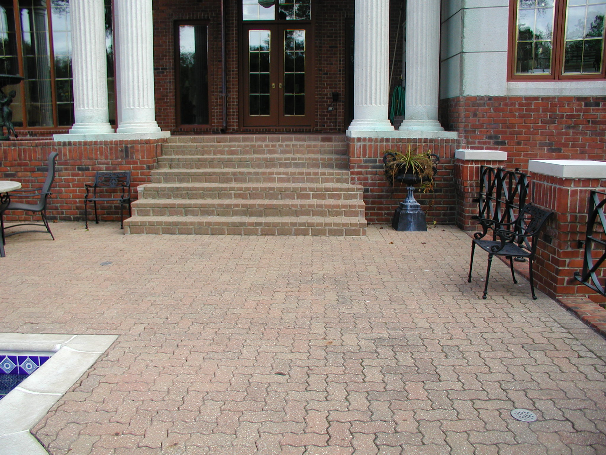Brick patio with wrought iron chairs, adjacent to an entrance with large columns and steps. No recognizable landmarks or historical buildings visible.